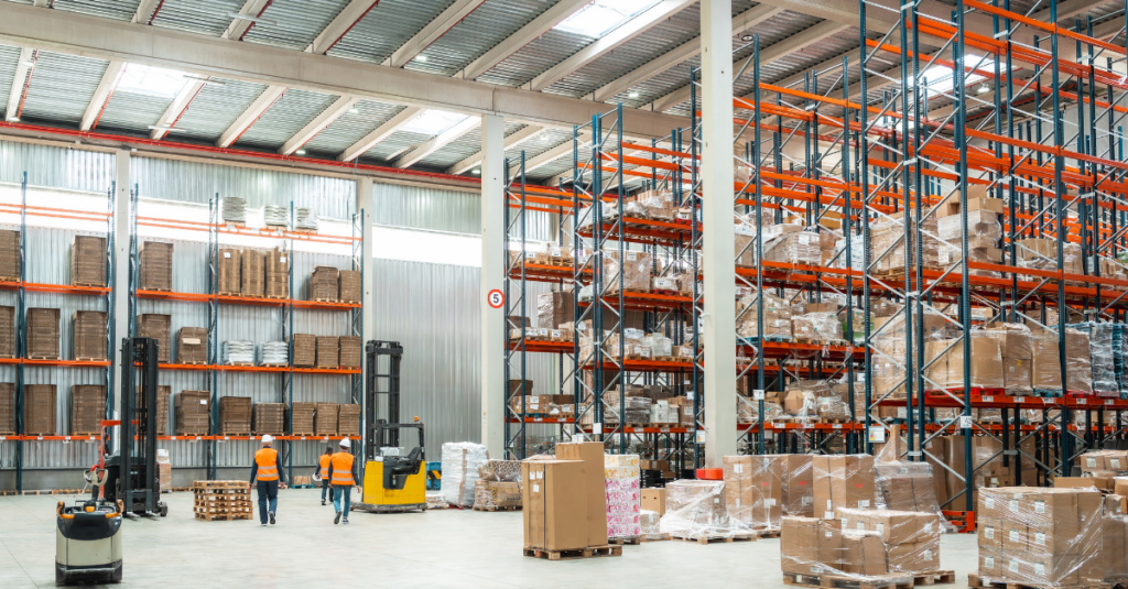 Large warehouse with tall pallet racking, forklifts, palletized inventory, and workers in high-visibility vests walking through storage area