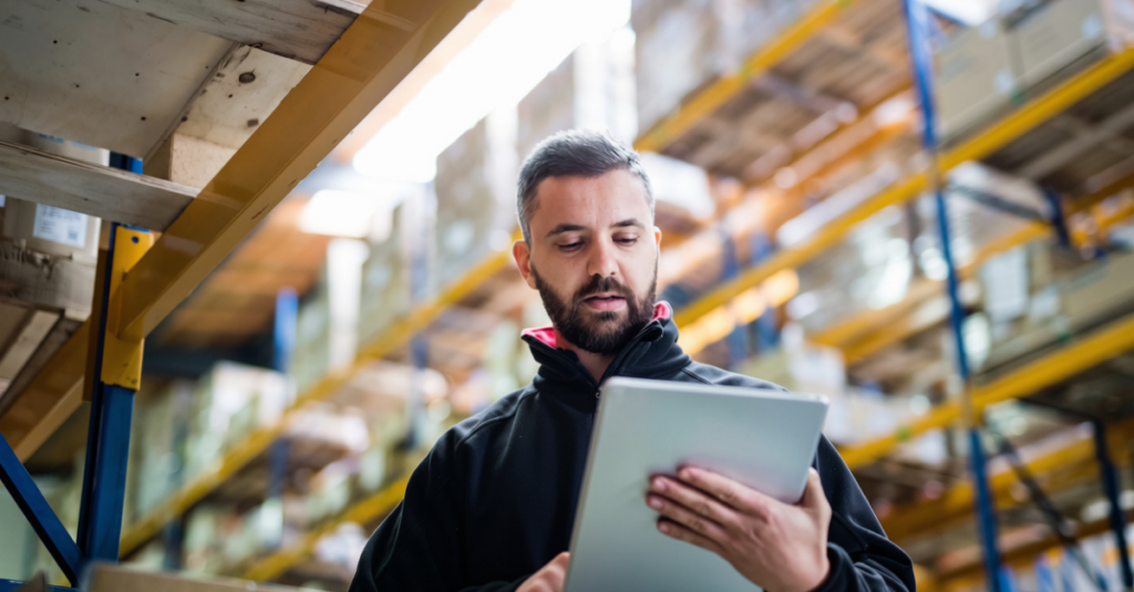 Person using tablet in warehouse aisle with pallet racking and boxed inventory stored on shelving