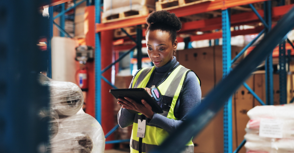 Warehouse worker in high-visibility vest using tablet beside palletized inventory and warehouse storage racks