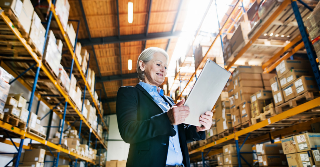 Person using tablet in warehouse aisle between tall storage racks stocked with boxes and palletized inventory