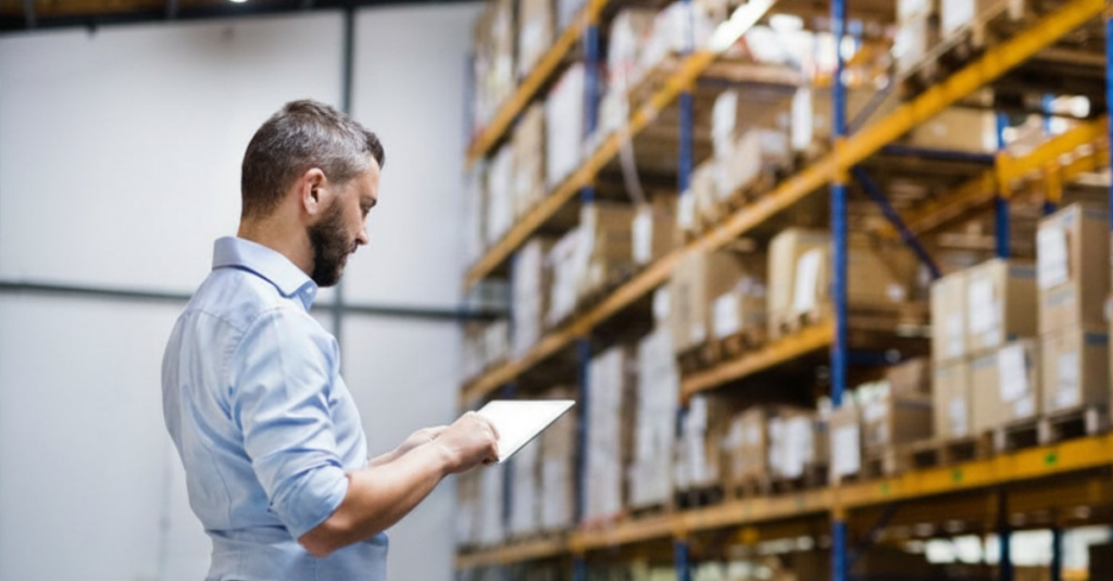 Person using tablet to review inventory in warehouse aisle with pallet racking and boxed inventory