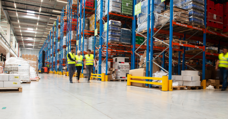 Warehouse aisle with tall pallet racking, palletized inventory, and workers in high-visibility vests walking through storage area