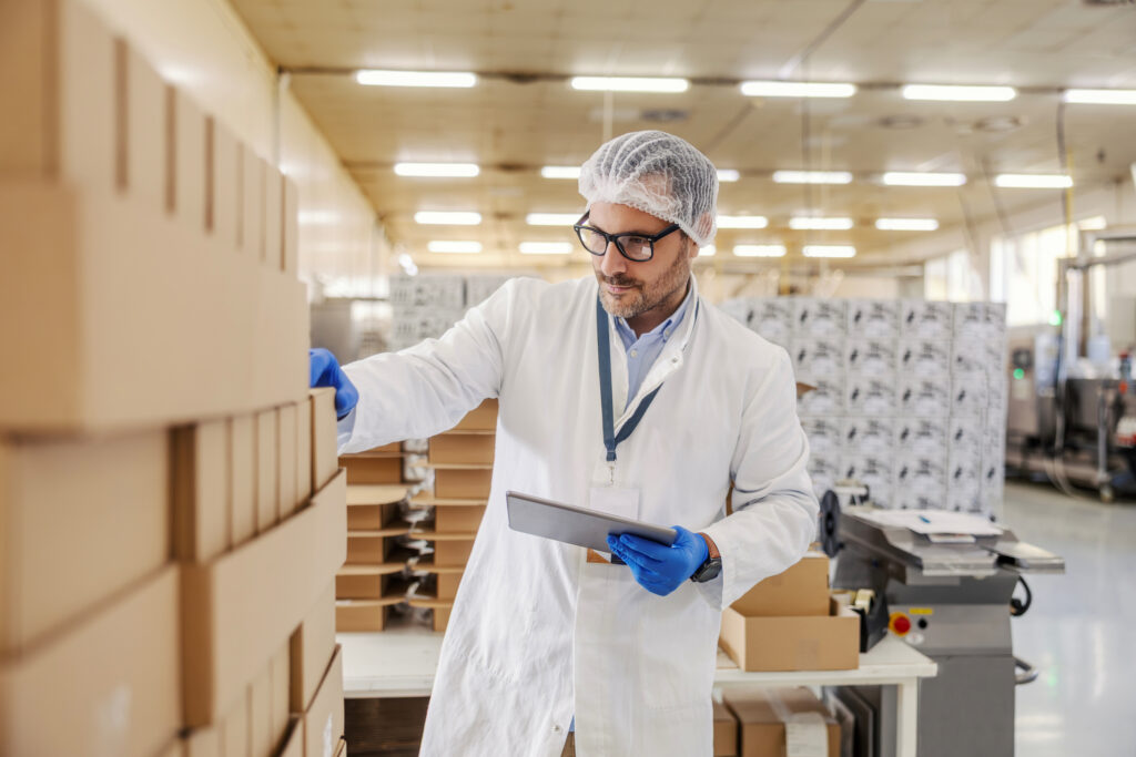 Worker in hairnet and lab coat using tablet while checking boxed inventory in regulated production facility