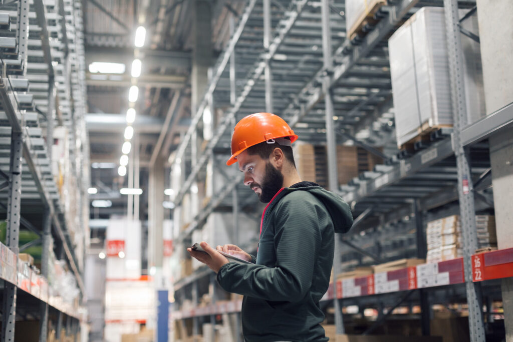 Warehouse worker using a tablet while conducting inventory checks in an aisle lined with pallet racks and stored goods.