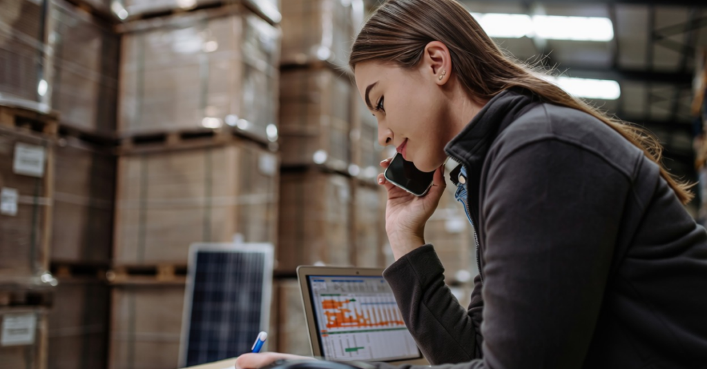 Person speaking on phone and taking notes beside laptop in warehouse with stacked pallets