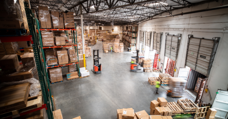 Large distribution center with palletized inventory, dock doors, forklifts, and workers moving freight through warehouse floor