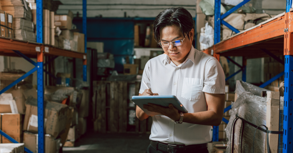 Person using tablet in warehouse aisle with storage racks and boxed inventory on shelving