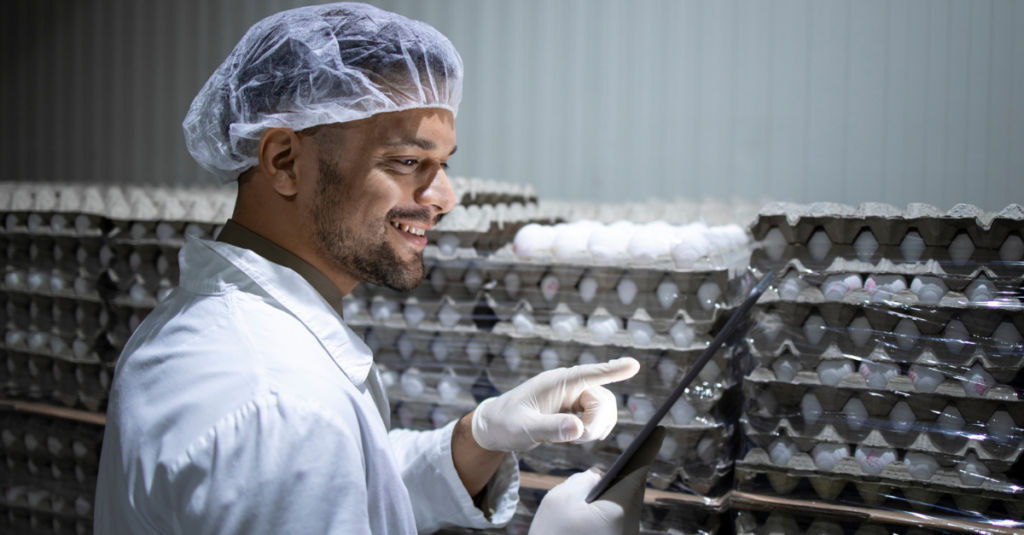 Person in lab coat, hairnet, and gloves using tablet beside stacked cartons of eggs in food production facility