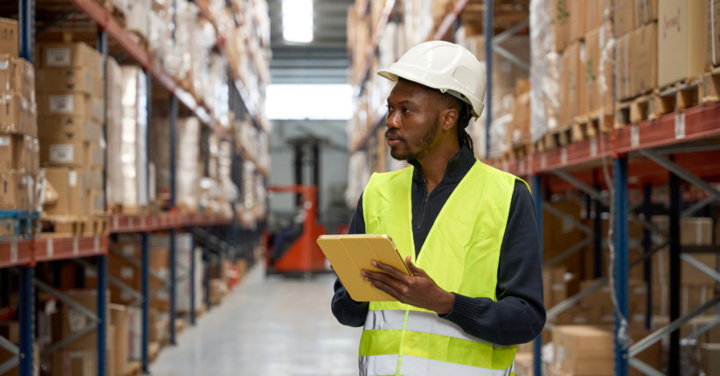 Warehouse worker wearing hard hat and high-visibility vest using tablet between racks of boxed inventory