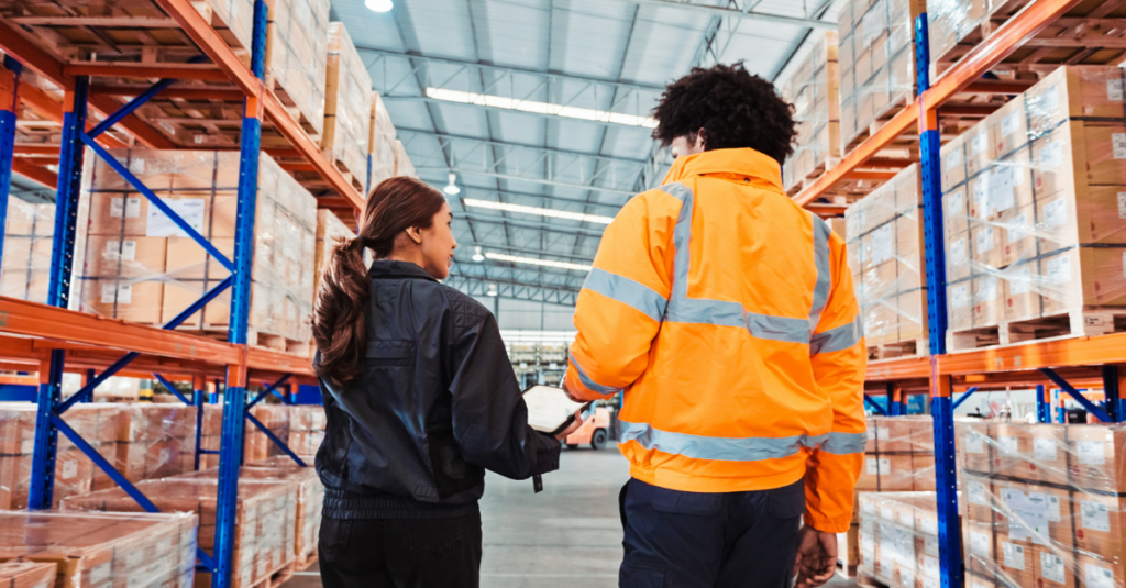 Two warehouse workers reviewing inventory information while walking through aisle with pallet racking and boxed inventory