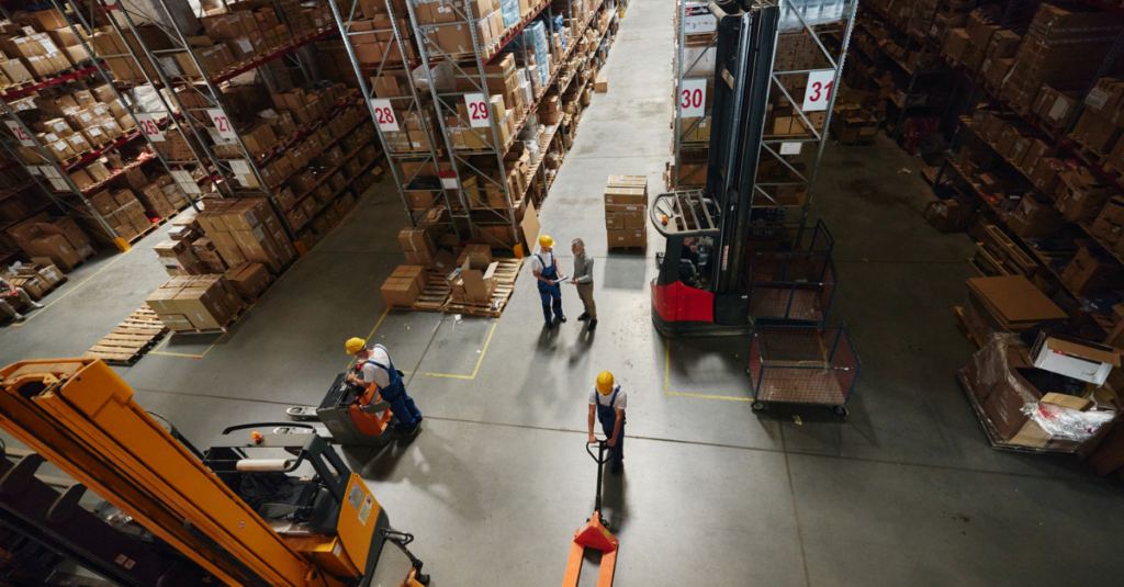 Overhead view of warehouse workers, forklifts, and pallet jacks moving inventory between storage aisles and palletized boxes