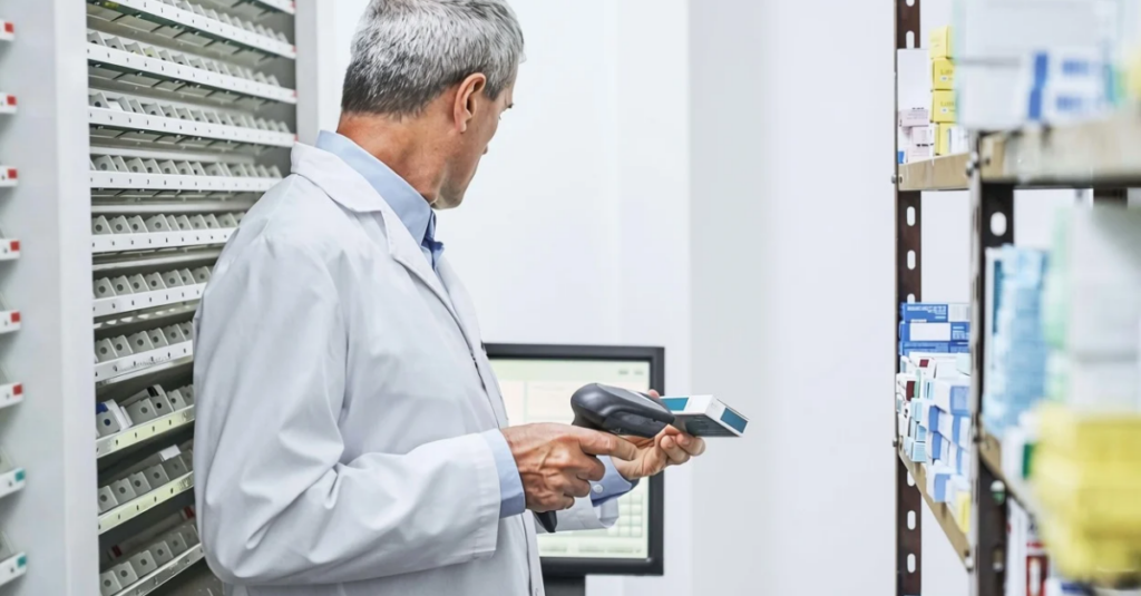 Person in lab coat using handheld barcode scanner to check medication inventory on pharmacy shelving