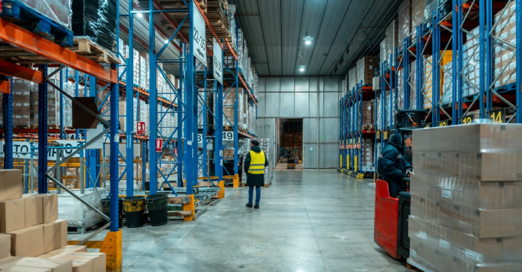 Cold storage warehouse aisle with palletized inventory on tall racking, worker in high-visibility vest, and forklift moving product